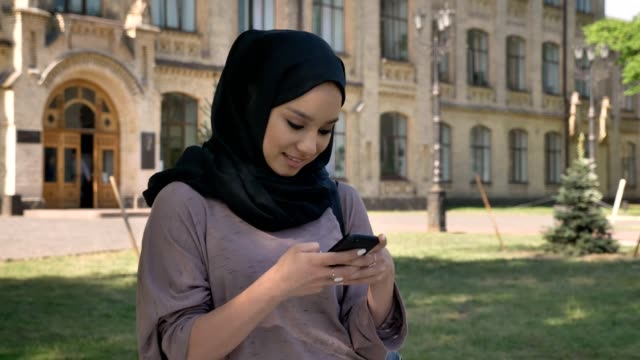 Young-sweet-muslim-girl-in-hijab-is-typing-message-on-smartphone-in-daytime-in-summer,-giggling,-building-on-background,-religiuos-concept,-communication-concept