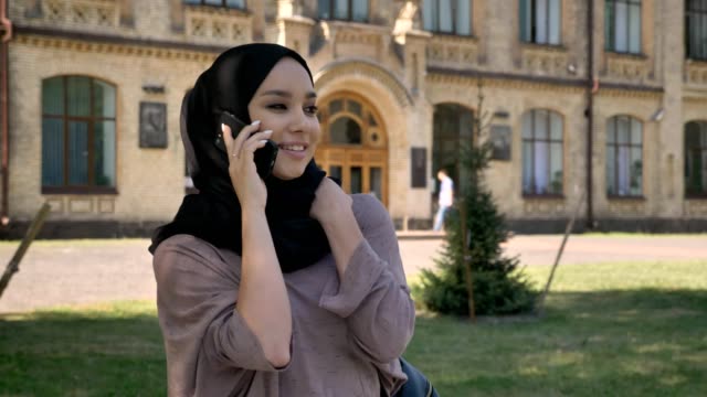 Young-sweet-muslim-girl-in-hijab-is-talking-on-phone-and-laughing-in-daytime-in-summer,-building-on-background,-religiuos-concept,-communication-concept