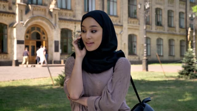 Young-sweet-muslim-girl-in-hijab-is-talking-on-phone-and-hand-up-in-daytime-in-summer,-building-on-background,-religiuos-concept,-communication-concept