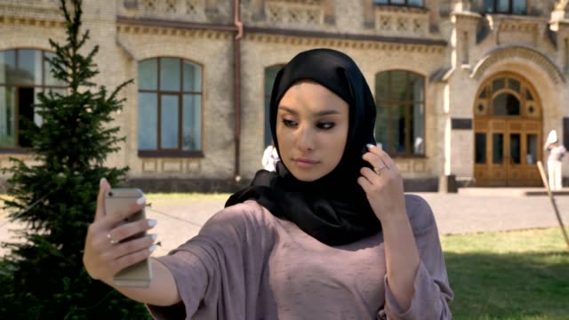 Young-beautiful-muslim-girl-in-hijab-is-making-selfie-on-her-smartphone,-showing-like-sign-in-daytime-in-summer,-smiling,-building-on-background,-religiuos-concept,-communication-concept