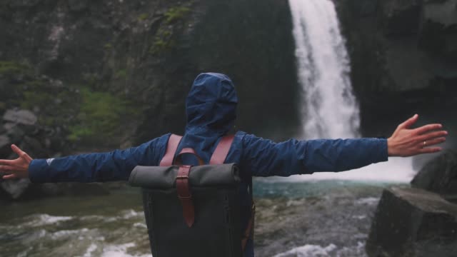 Back-view-shot-of-young-man-rising-hands-near-the-powerful-waterfall-in-Iceland,-cinematic-shot