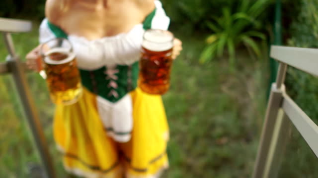 Sexy-Oktoberfest-waitress-carries-two-large-glasses-of-beer.-The-girl-walks-barefoot-on-the-grass-and-climbs-the-stairs.-Bavarian-traditional-costume,-erotic-neckline