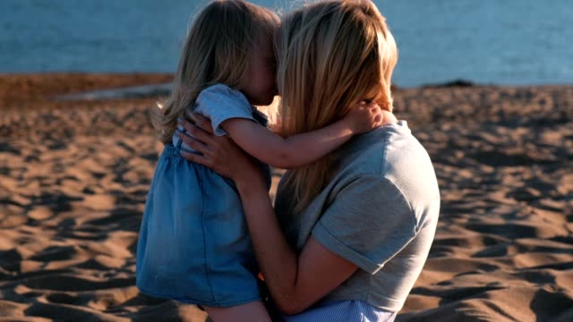Beautiful-blonde-mom-and-daughter-cuddling-on-the-beach-at-sunset.