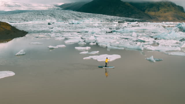 Toma-aérea-del-hombre-remando-de-pie-Junta-de-paleta-en-la-laguna-de-glaciar-con-icebergs-gigantes-en-Islandia