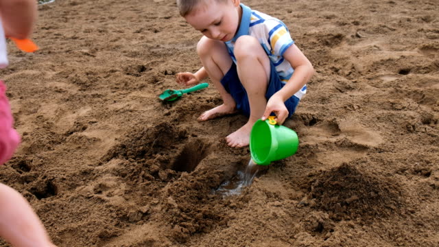Kleiner-Junge-gießt-Wasser-aus-einem-Eimer-in-ein-Loch-in-den-Sand.-Kinder-spielen-am-Strand