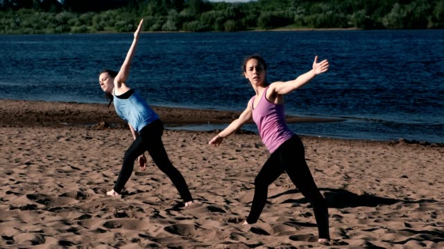 Zwei-Frau-Yoga-am-Sandstrand-am-Fluss-in-der-Stadt-zu-tun.-Blick-auf-die-schöne-Stadt-im-Sonnenaufgang.-Trikonasans-Utiha-darstellen.