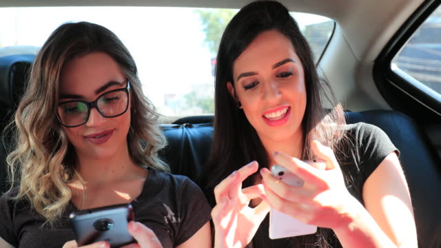 Candid-friends-holding-cellphones-in-the-back-seat-of-a-taxi-typing-and-sending-messages.-Girlfriends-browsing-the-internet