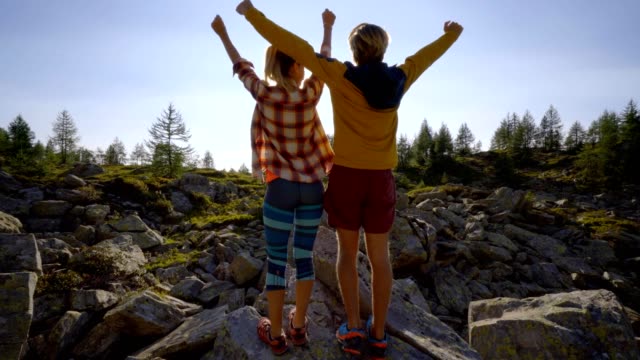 Young-couple-on-a-hike-surrounded-by-mountain-peaks-standing-arms-outstretched-in-front-of-the-sun,-Couple-hiking-arms-wide-open-freedom-and-achievement-concept