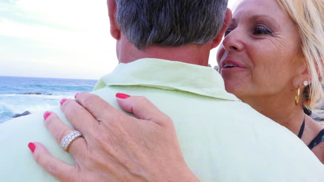 Close-up-of-an-older-woman-smiling-and-whispering-"I-love-you"-into-her-husband's-ear-at-the-beach
