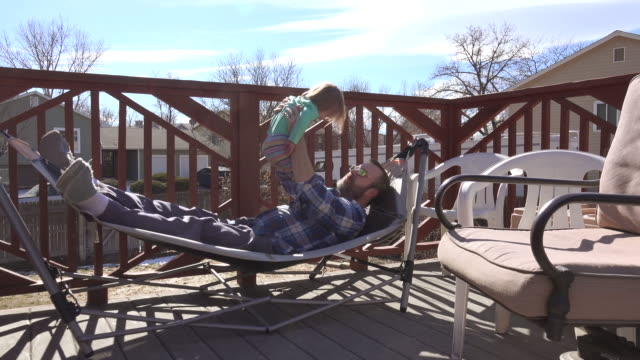 Vater-mit-Kleinkind-auf-Terrasse-Hängematte-im-Sommersonne-spielen.