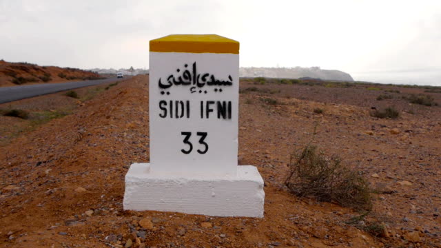 Nahaufnahme-der-Distanzschilder-nach-Sidi-infi-in-französischer-und-arabischer-Sprache-mit-Kutschen-im-Hintergrund.-Marokko