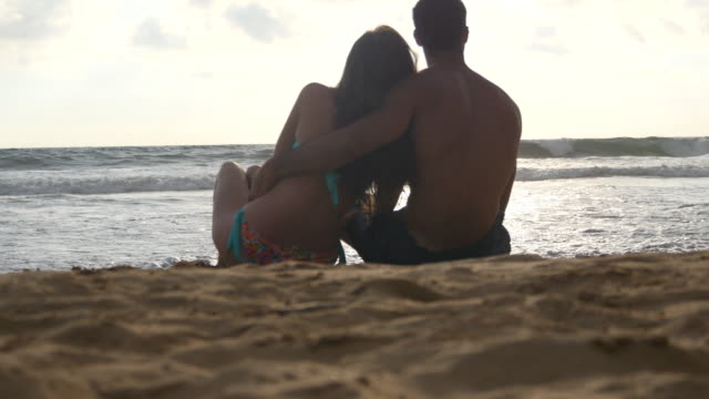 Young-romantic-couple-is-enjoying-beautiful-view-sitting-on-the-beach-and-hugging.-A-woman-and-a-man-sits-together-in-the-sand-on-the-seashore,-admiring-the-ocean-and-landscapes.-Close-Up