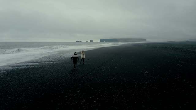 Copter-vuela-después-de-joven-pareja-en-la-orilla-del-mar-en-Jersey-islandés.-Negro-playa-volcánica-en-Islandia