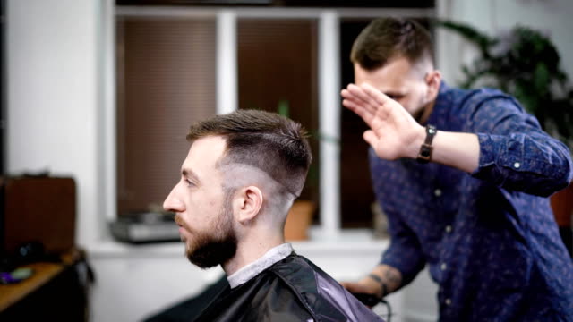 Young-barber-standing-and-making-stylish-haircut-of-attractive-man-with-clipper-in-barbershop.-Bearded-man-is-sitting-on-the-chair-with-black-protective-cape-against-the-mirror-and-looking-at-himself