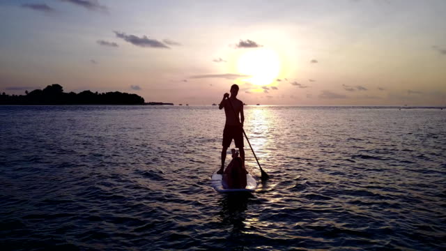 v04105-fliegenden-Drohne-Luftaufnahme-der-Malediven-weißen-Sandstrand-2-Personen-junges-Paar-Mann-Frau-Paddleboard-Rudern-Sonnenuntergang-Sonnenaufgang-auf-sonnigen-tropischen-Inselparadies-mit-Aqua-blau-Himmel-Meer-Wasser-Ozean-4k