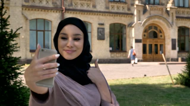 Young-beautiful-muslim-girl-in-hijab-is-making-selfie-on-her-smartphone,-showing-like-sign-in-daytime-in-summer,-building-on-background,-religiuos-concept,-communication-concept