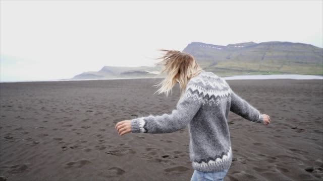 Brazos-de-la-joven-extendidos-por-el-pie-de-mar-en-la-playa-de-arena-negra,-de-pelo-en-el-viento-Islandia---mujer-correr-juguetón-disfrutar-naturaleza-y-libertad---lenta