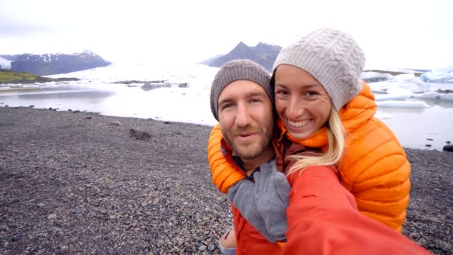 4K-Video-of-young-couple-having-fun-taking-selfie-by-the-glacier-lake-at-Jokulsarlon-lagoon-in-Iceland.-Two-people-travel-love-romance-sharing-togetherness-communication-concept.-Springtime,-cold-temperatures