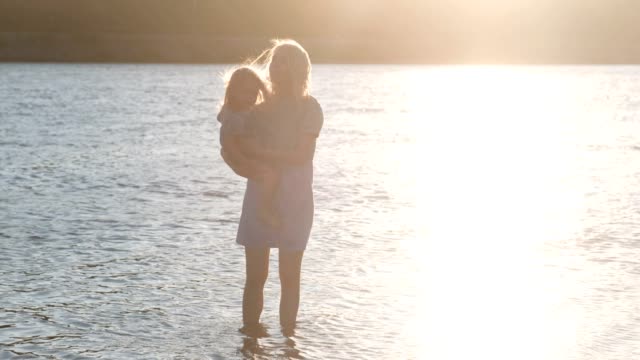 Beautiful-blonde-mother-and-daughter-walk-along-the-river-on-the-water-at-sunset.