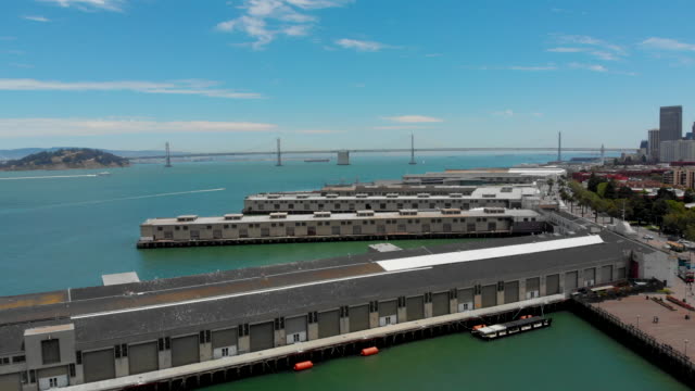Drohne-geschossen-aufsteigend-über-eine-Gruppe-von-kommerziellen-Docks-in-San-Francisco-mit-Boote,-blauer-Himmel,-Wasser-und-der-San-Francisco-Oakland-Bay-Bridge-im-Blick
