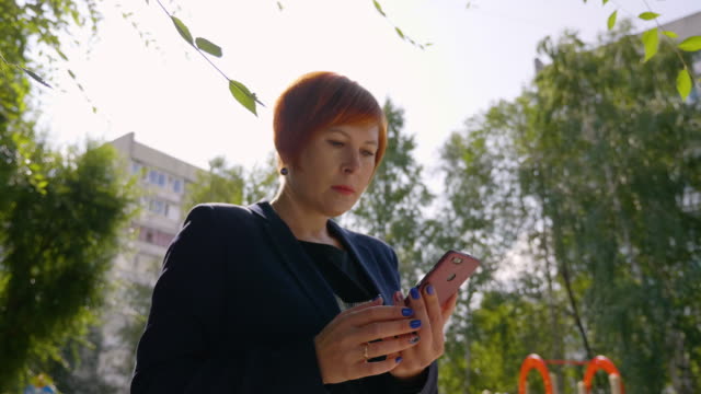 Serious-woman-with-red-hair-using-smartphone-outdoors-with-backlit,-low-angle