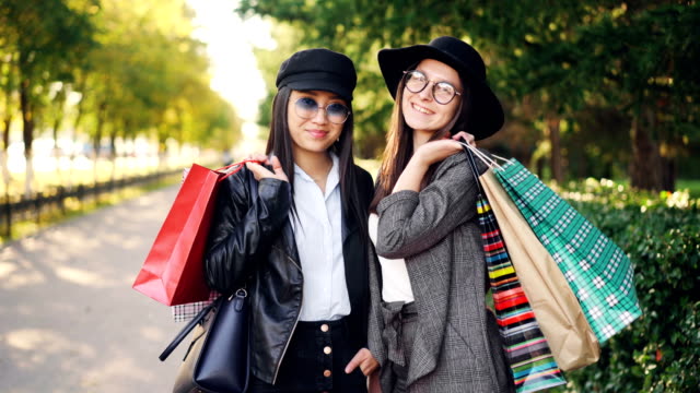 Portrait-of-beautiful-young-women-shoppers-standing-in-the-street-holding-paper-bags-and-smiling-looking-at-camera.-Youth-lifestyle-and-consumerism-concept.