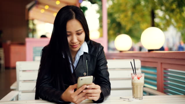 Modern-Asian-woman-is-using-smartphone-texting-friends-sitting-in-street-cafe-on-windy-autumn-day-and-smiling.-Technology,-communication-and-people-concept.