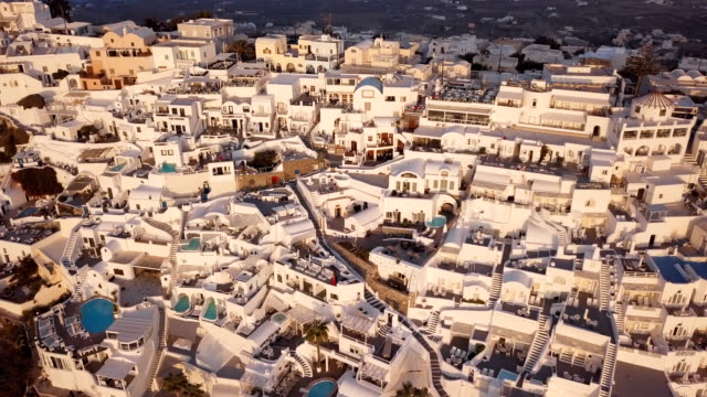 Vuelo-sobre-villas-blancas-terrazas-tradicionales-ciudad-de-Fira-(Thira)-al-atardecer,-Santorini,-Grecia