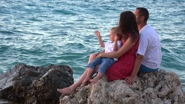 Young-parents-and-happy-child-sitting-together,-rocky-seashore-waves