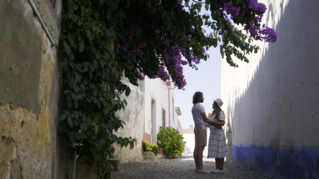 Couple-kissing-between-old-buildings-near-shrub-with-blooms