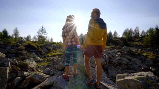Young-couple-on-a-hike-surrounded-by-mountain-peaks-standing-arms-outstretched-in-front-of-the-sun,-Couple-hiking-arms-wide-open-freedom-and-achievement-concept