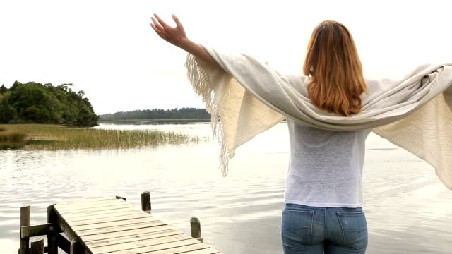 Young-woman-relaxes-on-lake-pier,-stands-arms-outstretched