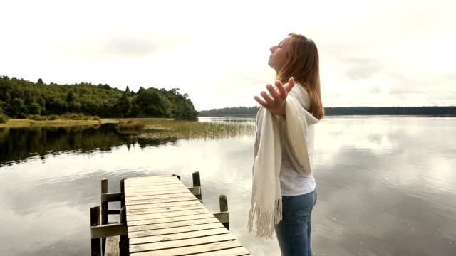 Young-woman-relaxes-on-lake-pier,-stands-arms-outstretched