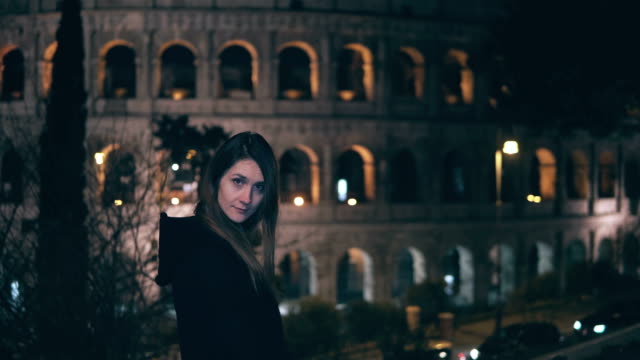 Retrato-de-mujer-morena-joven-de-pie-cerca-de-Coliseo-de-Roma,-Italia-en-la-noche.-Niña-da-vuelta-y-mira-a-cámara