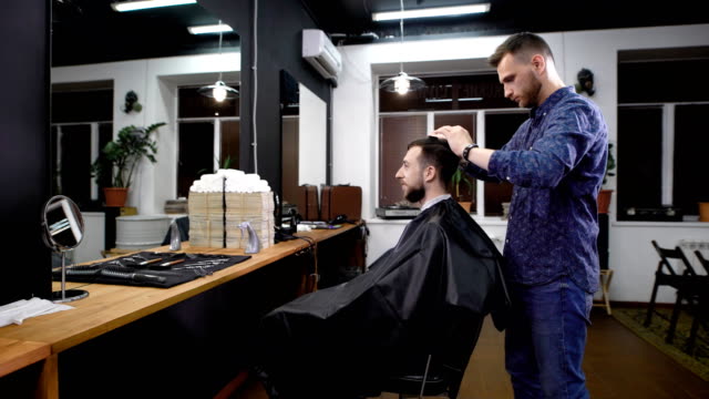 Jungen-Barbier-in-Freizeitkleidung-stehend-und-Client-vor-dem-Schneiden-der-Haares-kämmen.-Lächelnder-Mann-sitzen-auf-dem-Stuhl-mit-schwarzen-Frisiermantel-bedeckt-und-mit-Blick-auf-den-Spiegel-gegenüber-im-Frisörsalon
