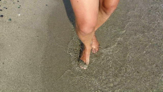 Men-barefoot-legs-walking-along-sea-shore-shallow-water-with-waves-splashing.-4k-trekking-steadicam--front-view-pov-shot