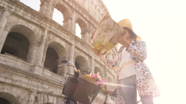 Beautiful-young-woman-in-colorful-fashion-dress-with-bike-reading-map-in-front-of-colosseum-in-Rome-at-sunset-with-happy-attractive-tourist-girl-with-straw-hat-looking-for-directions-ground-shot-dolly-moving-camera