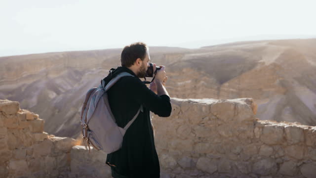 Male-photographer-walks-along-ancient-Israel-ruins.-Road-to-Masada-fortress.-Young-man-with-backpack-takes-photos.-4K