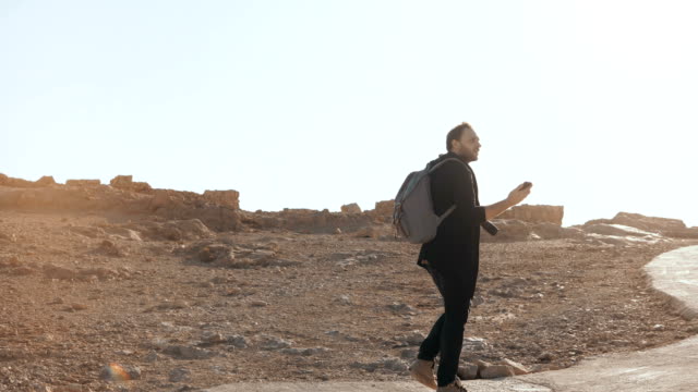 European-man-walks-to-ancient-ruins,-looks-around.-Relaxed-male-tourist-makes-phone-videos-on-desert-road.-Israel-4K
