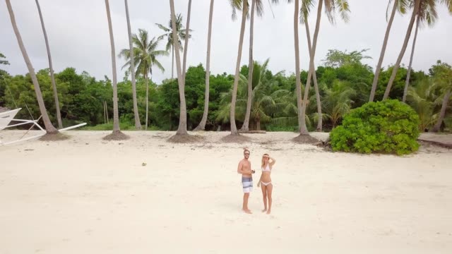 Young-couple-playing-with-drone,-waving-hands-to-the-flying-helicopter.-People-fun-new-technology-vacations-concept.-Shot-on-tropical-beach-in-the-Philippines,-4K-resolution
