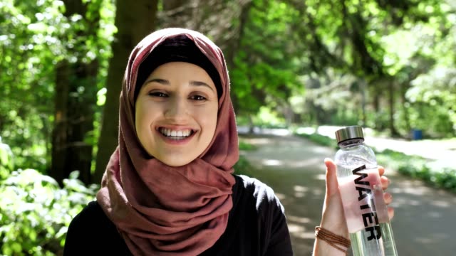 Retrato-de-una-linda-chica-joven-en-un-hiyab-con-una-botella-de-agua-en-sus-manos,-mostrando-como-señal,-sonriendo,-mirando-a-la-cámara,-parque-al-fondo.-50-fps