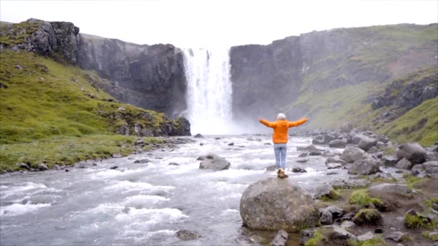 Junge-Frau-vor-dem-herrlichen-Wasserfall-in-Island-ausgestreckten-Armen.-Die-Leute-reisen-Exploration-Konzept-Zeitlupe