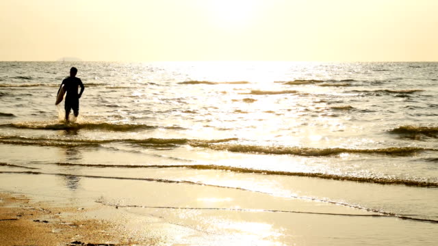 4-K.-Silhouette-des-jungen-glücklich-Surf-Mann-läuft-mit-langen-Surfbretter-bei-Sonnenuntergang-am-tropischen-Strand.-Surfer-am-Strand-im-Meer-bei-Sonnenuntergang-mit-schönen-Licht-mit-Sonnenlicht-flare.
