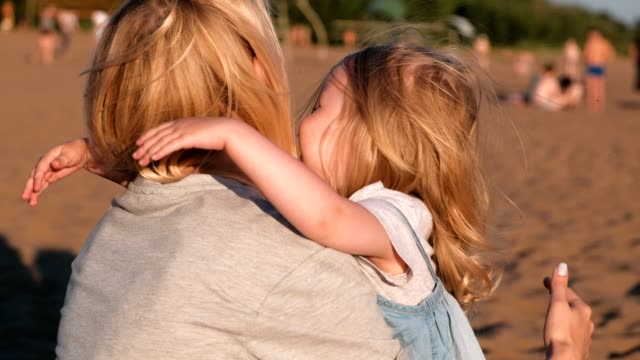 Beautiful-blonde-mom-and-daughter-cuddling-on-the-beach-at-sunset.