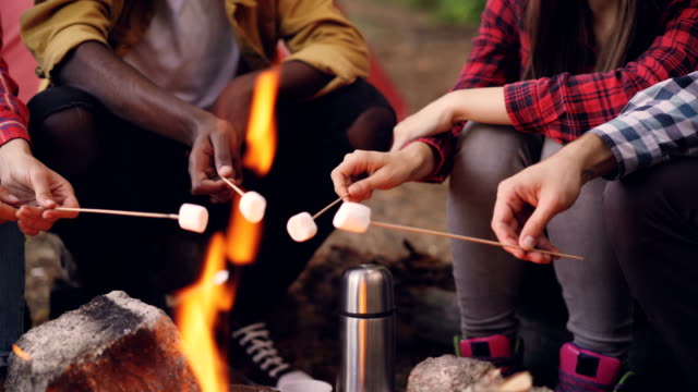 Multiethnic-group-of-tourists-is-cooking-food-on-fire-warming-marshmallow-on-sticks-resting-around-campfire-during-hike.-People's-arms-and-legs-are-visible.