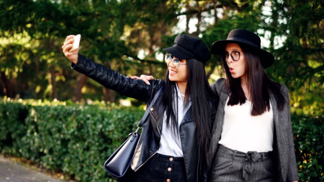 Fröhliche-junge-Damen-Shopaholics-nehmen-Selfie-auf-der-Straße-halten-Taschen-und-lustige-Grimassen.-Frauen-tragen-elegante-Kleidung-und-Brille.