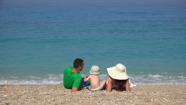 Family,-young-parents-and-little-child-resting-on-beach,-turquoise-sea-waving