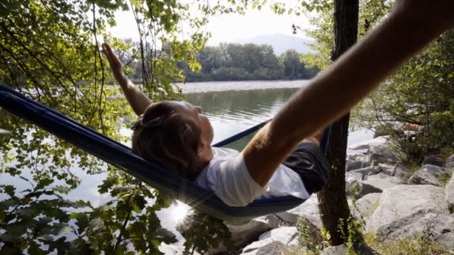 Young-woman-relaxing-on-hammock-by-the-river,-hands-behind-head-enjoying-serene-green-environment.-People-travel-relax-concept-in-vacations