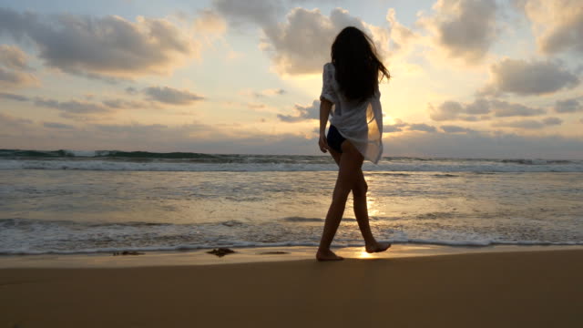 Happy-woman-walking-on-the-beach-of-the-ocean-and-spraying-the-water-with-her-feet.-Young-beautiful-girl-enjoying-life-and-having-fun-at-sea-shore.-Summer-vacation.-Sunset-landscape-at-background