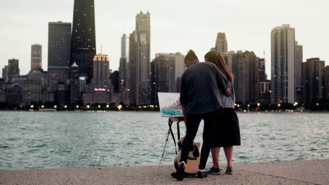 Kreatives-junges-Paar-am-Ufer-des-Michigan-Sees,-Chicago,-Amerika.-Frau,-Zeichnung,-Mann,-hält-die-Gitarre-Abend